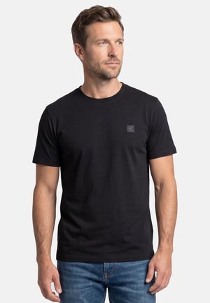 Mand i en ensfarvet sort kortærmet t-shirt og blå jeans, der ser let mod højre mod en ensartet lys grå baggrund.