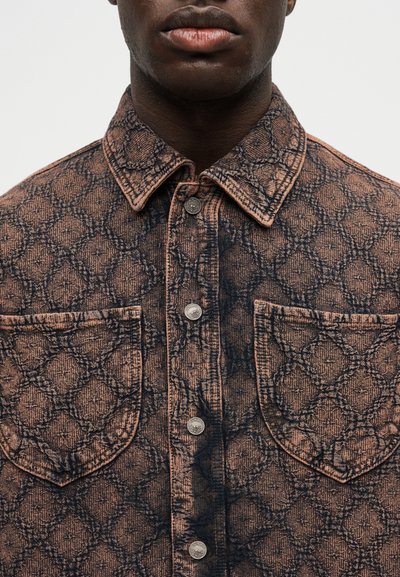 Άνδρας με καφέ πουκάμισο με κουμπιά, σχέδιο με δύο τσέπες στο στήθος και γεωμετρικό, υφασμάτινο μοτίβο.