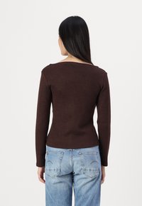 Haut marron à manches longues avec un col rond, coupe slim et texture douce, associé à un jean bleu clair avec des détails de poches arrière surpiqués.