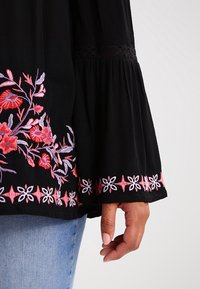 Blusa nera con ricami floreali in rosso e rosa sulla parte anteriore e sui bordi delle maniche. Maniche a campana plissettate con dettagli in pizzo alle spalle.