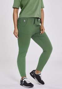 Green cotton joggers with a drawstring waist, side pockets, and a fitted design. Worn with black sneakers featuring white accents.