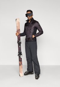 Melna pūkaina jakas ar rāvējslēdzēju, tumši bikses, tur rokā slēpi. Slēpe ir ar brūnu un purpura dizainu ar stiprinājumiem. Valkā melnas aizsargbrilles.