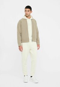 Beige kordjakke, creme hættebluse og hvide joggers. Glat tekstur med lynlås. Modellen har hvide sneakers på. Enkelt design.