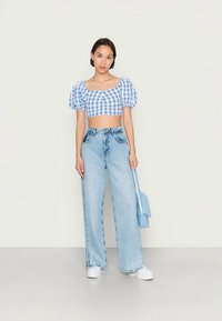 Jovem mulher de pé, vestindo um top cropped de manga bufante em azul e branco xadrez, calças de cintura alta em azul claro e largas, ténis brancos, segurando uma bolsa azul.