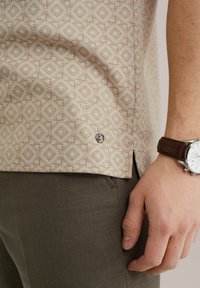 Camisa de manga corta en beige claro con un patrón de diamantes. El dobladillo presenta una pequeña etiqueta de logo. Combinada con pantalones en verde oliva.