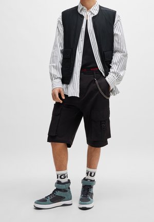 Gilet senza maniche nero sopra una camicia a righe con bottoni, pantaloni cargo neri e sneakers turchesi con suole bianche. Cintura rossa con marchio e catena come accessorio.