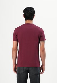 Mann mit kurzen dunklen Haaren, der ein einfaches burgunderfarbenes T-Shirt und schwarze Jeans trägt, steht mit dem Rücken zu einem weißen Hintergrund.