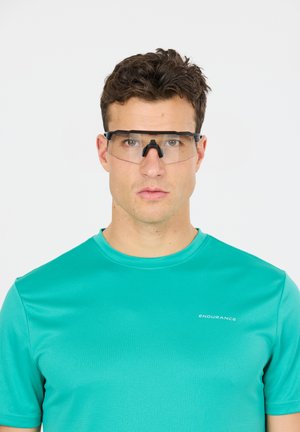 Homme portant des lunettes de sport transparentes et un maillot athlétique turquoise avec le logo « Endurance », face à face sur un fond blanc uni.