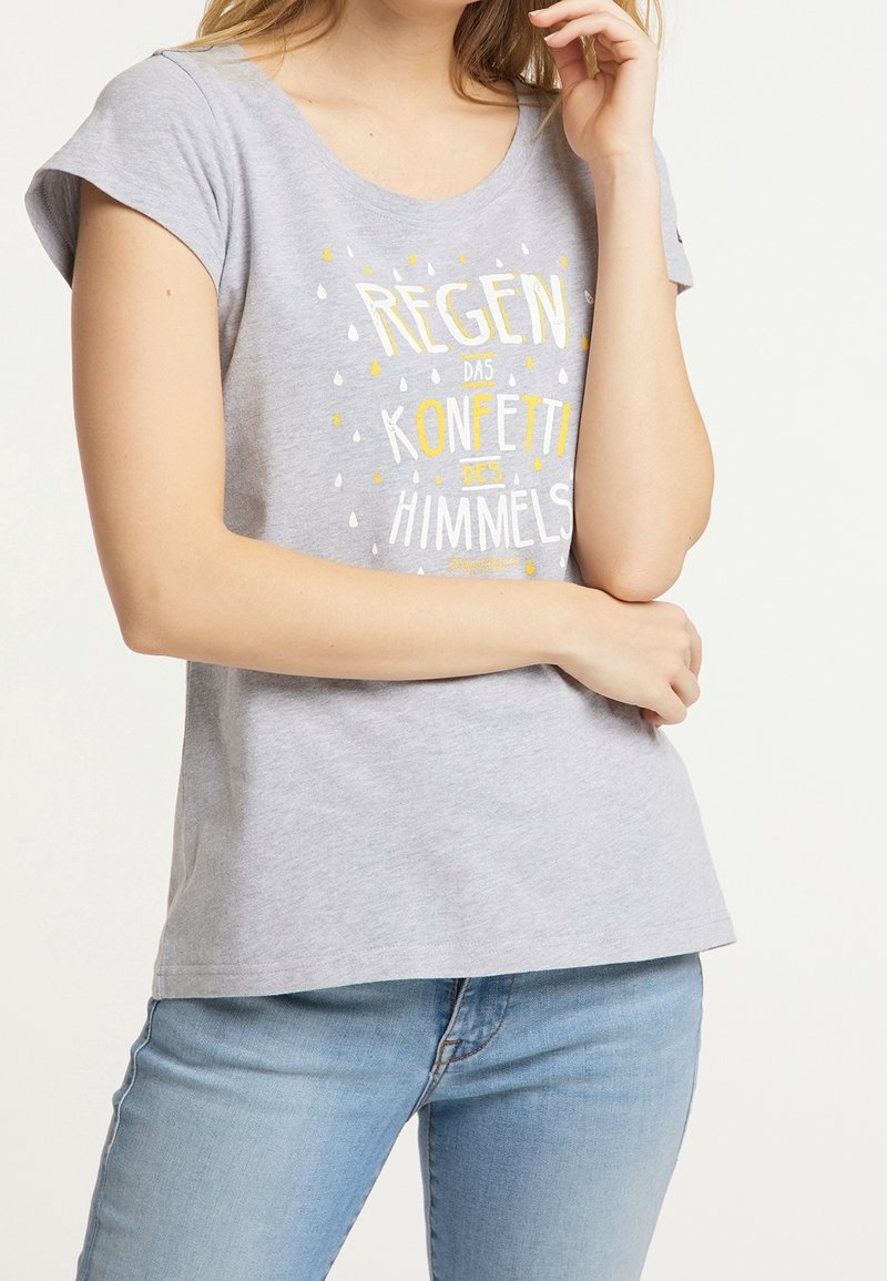 Femme portant un t-shirt gris avec un texte jaune et blanc et un motif de gouttes de pluie, associé à un jean bleu clair, posant les bras croisés.