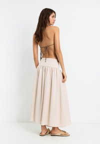 Beige maxi skirt with a high, elastic waistband and a flowing design. The model wears a backless top with adjustable ties.