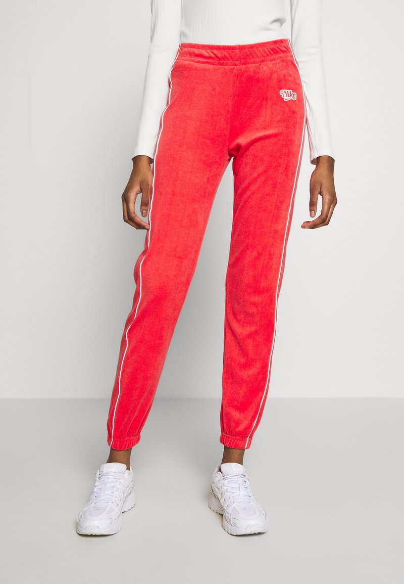 Röda velour joggers med vita sidostrick, elastisk midja och muddar vid anklarna. Litet Nike-logotyp på vänster lår.