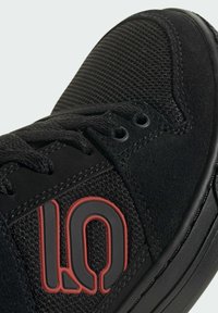 Primo piano di una sneaker nera con tessuto strutturato, pannelli in camoscio, lacci neri e un logo "10" in rilievo contornato di rosso sul lato.