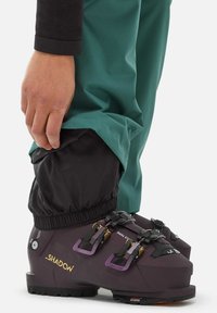 Skischuhe in Dunkelpurpur mit gelben und lila Schnallen, getragen mit schwarzer und grüner Outerwear. Verfügt über ein strukturiertes Material und eine sichere Passform.