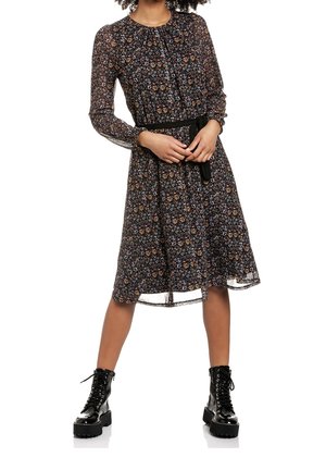 Nainen, joka pukeutunut mustaan, kukalliseen polvipituiseen mekkoon, jossa on pitkät hihat ja musta vyö, yhdistettynä mustiin kiiltäviin nyörikengillä varustettuihin paksupohjaisiin saappaisiin.