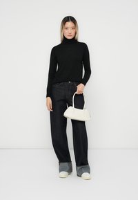 Pull à col roulé noir, jeans en denim sombre à jambes larges, accents gris aux poignets, sac à main blanc avec des anses arrondies, baskets blanches.
