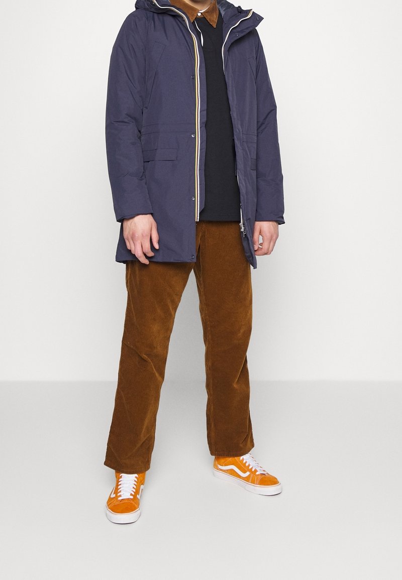 Personne portant une veste à capuche bleu marine, une chemise noire, un pantalon en velours côtelé marron et des baskets orange vif et blanches, debout sur un fond blanc.