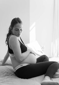 Zwarte sportbeha met brede banden en logo, zwarte legging, zittende pose op een licht gestructureerde deken, die een duidelijk afgeronde buik benadrukt.