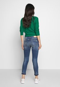 Jersey verde con mangas enrolladas, combinado con jeans ajustados de lavado claro que tienen bolsillos traseros, usados con zapatillas blancas.