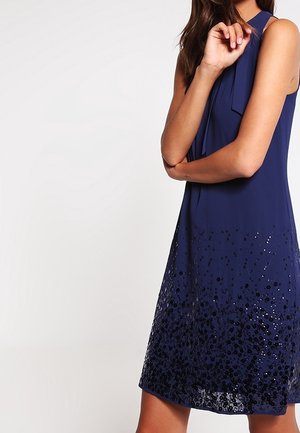 Granatowa sukienka bez rękawów z kokardą przy dekolcie i cekinowym kwiatowym wzorem wzdłuż dolnego brzegu, noszona przez osobę ze skrzyżowanymi ramionami.