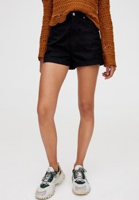 Shorts in denim neri con vita alta, risvolti e ampie tasche frontali, abbinati a scarpe da ginnastica bianche e verdi.