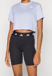 Camiseta recortada de color púrpura claro con mangas cortas y logo blanco; pantalones cortos negros de cintura alta con cinturilla estampada. Tejido suave de mezcla de algodón.