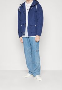 Giacca a vento blu navy con cappuccio e lacci bianchi, grandi tasche e polsini a costine, abbinata a jeans a gamba larga blu chiaro e sneakers bianche.