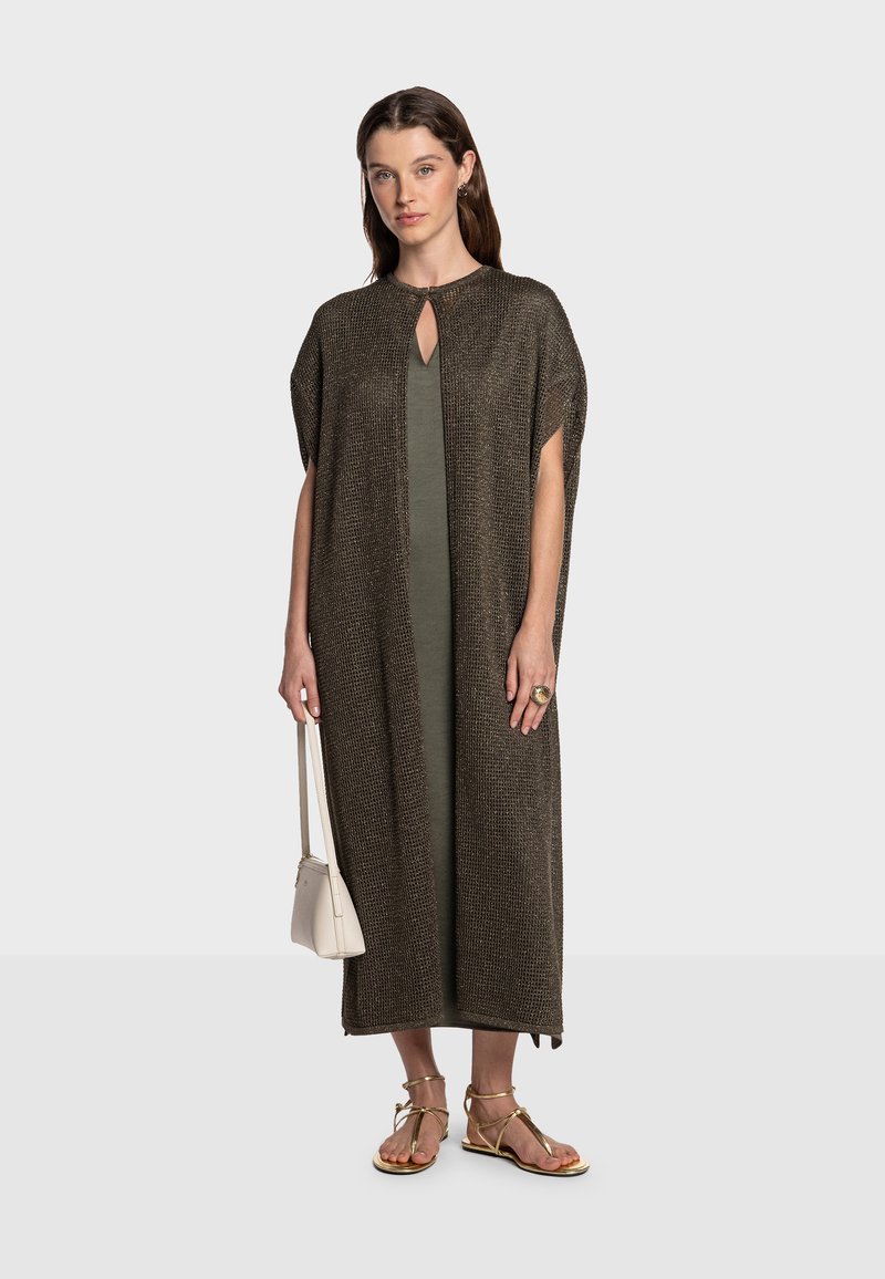 Kaftán marrón tejido con escote de ojo de cerradura, combinado con un vestido verde. La modelo sostiene una bolsa blanca y lleva sandalias doradas.