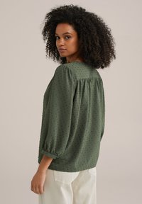 Blusa verde con tessuto a trama, maniche a palloncino e piccoli puntini in rilievo. Il design sul retro presenta un dettaglio arricciato, indossata con pantaloni di colore chiaro.
