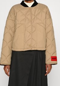 Person iført en lysbrun, kviltet jakke med tryknapper, sort krave, rød HUGO-logo patch på ærmet og en sort plisseret nederdel.