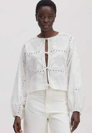 Vrouw draagt witte geborduurde blouse met strikjes aan de voorkant en lange opgeblazen mouwen, gecombineerd met witte broek, op een lichte achtergrond.