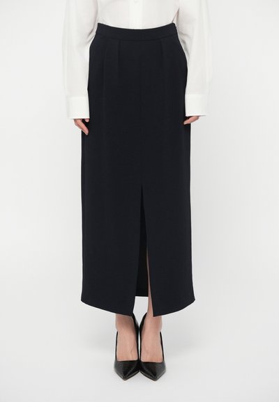 Femme portant une jupe midi noire avec une fente frontale, associée à une chemise blanche à manches longues et des escarpins noirs à bout pointu.