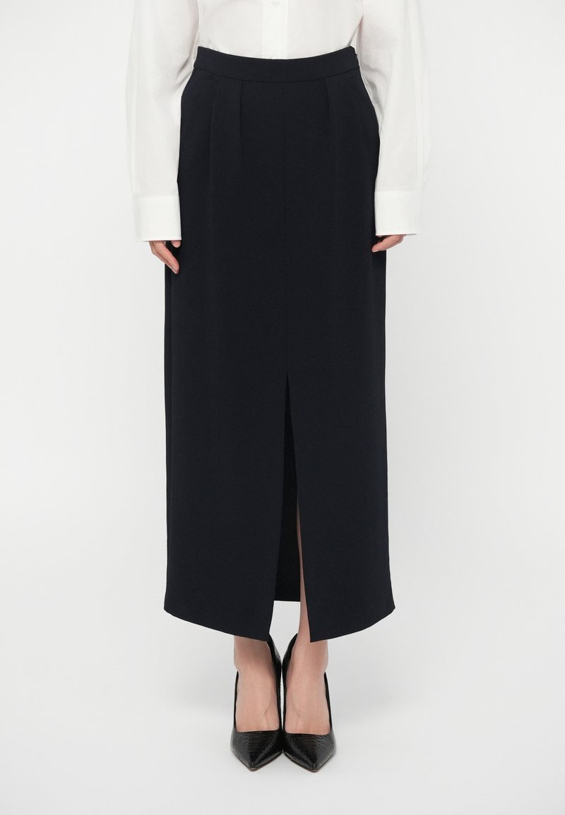 Γυναίκα που φοράει μια μαύρη μίντι φούστα με μπροστινή σκίτσο, συνδυασμένη με ένα λευκό πουκάμισο μακρυμάνικο και μαύρα παπούτσια με μύτη.