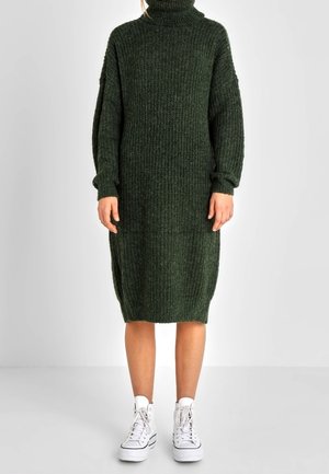 Knielanges, dunkelgrünes, geripptes Strickkleid mit Rollkragen, getragen mit weißen High-Top-Sneakers, Arme entspannt an den Seiten.