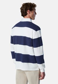 Mann in langärmligem Rugby-Shirt mit breiten blau-weißen horizontalen Streifen, beigen Hosen, steht mit Blick nach rechts vor einem weißen Hintergrund.