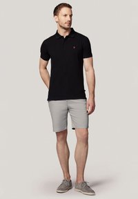 Czarna koszulka polo z krótkim rękawem i czerwonym logo, w połączeniu z jasnoszarymi szortami. Model ma na sobie szare buty, stoi na neutralnym tle.