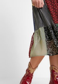 Robe en patchwork avec divers motifs en rouge, noir, vert et bleu, dotée d'un ourlet évasé ; associée à des bottes de cowboy rouges.