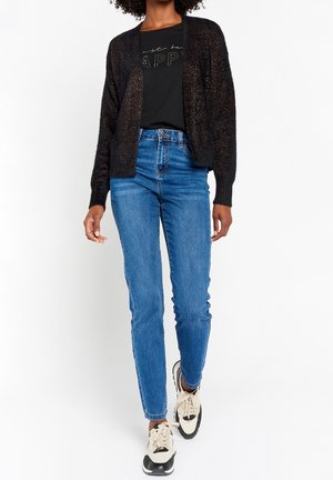 Person trägt eine schwarze, strukturierte Strickjacke, ein schwarzes T-Shirt mit der Aufschrift "just be happy", blaue Jeans und weiße-schwarze Sneakers und geht nach vorn.