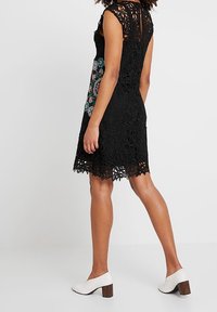 Robe en dentelle noire avec un motif floral, sans manches, ourlet festonné et dos en trou de serrure. Associée à des chaussures à talons carrés blanches.