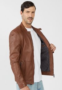 Braune Lederjacke mit Reißverschluss, gerippten Schulterdetails und schwarzem gemustertem Futter. Das Modell trägt ein weißes T-Shirt darunter.