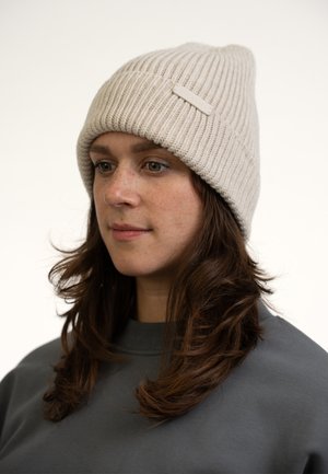Bonnet en tricot beige avec une texture côtelée. Présente une petite étiquette en relief à l'avant. Porté avec un sweat-shirt gris foncé.
