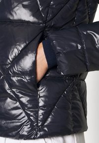 Marinblå puffjacka med en glansig yta, som har en diamantquiltad struktur och kontrasterande ribbade ärmslut. Sidfickdetalj synlig.