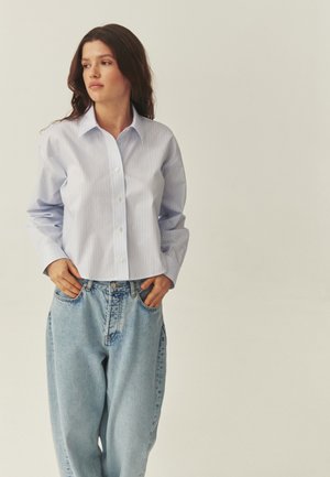 Młoda kobieta z długimi ciemnymi włosami, ubrana w jasnoniebieską koszulę w paski oraz luźne jasnoniebieskie dżinsy, ręce w kieszeniach, patrzy w lewo.