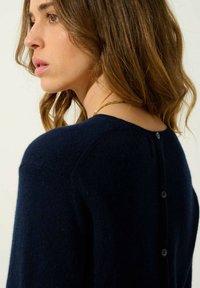 Granatowy sweter wykonany z miękkiego materiału, z okrągłym dekoltem i detalami z guzikami na plecach. Akcentowany luźnym krojem.