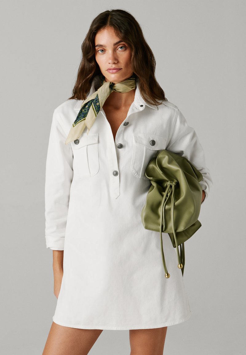 Ženska z dolgimi rjavimi lasmi, oblečena v belo obleko z gumbi in vzorčastim rutkim okoli vratu, ki drži veliko olivno zeleno torbo z vrvico.