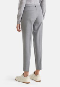 Osoba w jasnoszarych dopasowanych spodniach i beżowych sneakersach, stojąca na białym tle z rękami po bokach.