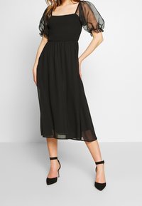 Schwarzes Kleid mit einer enganliegenden Raffung im Oberteil, durchsichtigen Puffärmeln und einem fließenden Rock. Kombiniert mit schwarzen Stiletto-Absätzen. Sanfte Stofftextur.