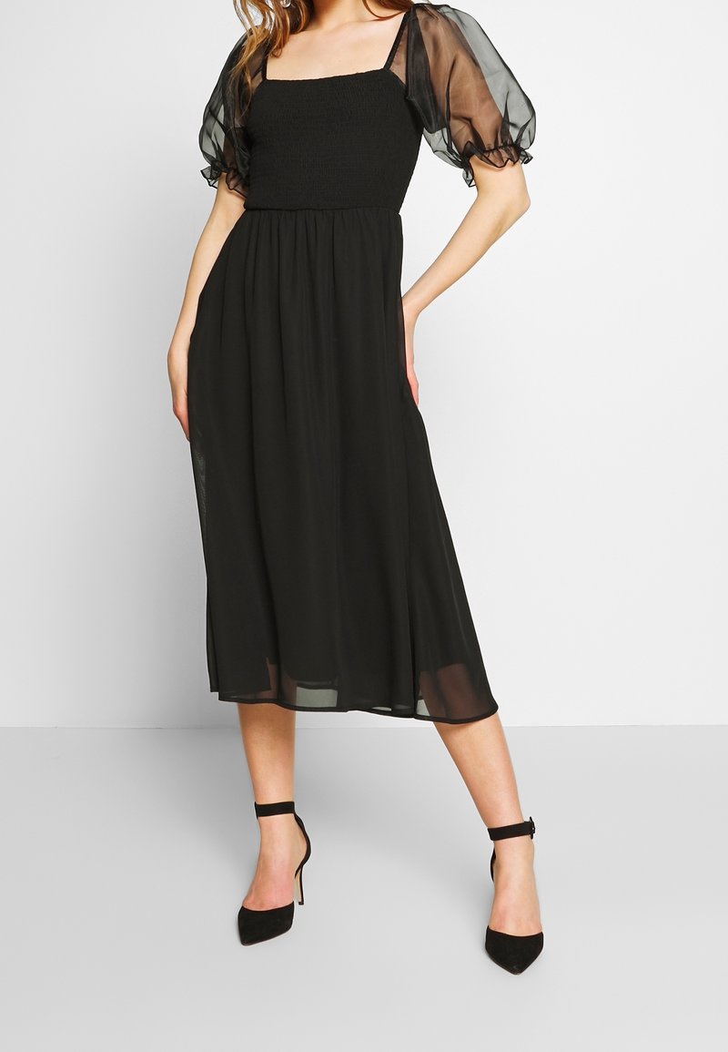 Schwarzes Kleid mit einer enganliegenden Raffung im Oberteil, durchsichtigen Puffärmeln und einem fließenden Rock. Kombiniert mit schwarzen Stiletto-Absätzen. Sanfte Stofftextur.