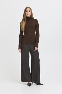 Femme debout en pantalon gris à jambes larges, pull à col roulé marron et talons noirs pointus sur fond blanc.