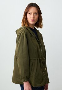 Giacca a vento verde oliva con cappuccio, vita regolabile, polsini elasticizzati, chiusura con zip frontale e due tasche laterali. Tessuto liscio e leggero.