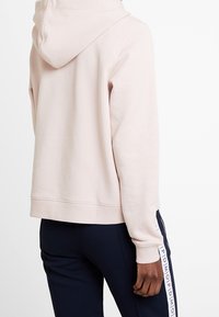 Sudadera con capucha rosa claro de textura suave, con capucha ajustable mediante cordón y puños acanalados, combinada con pantalones azul marino oscuro que muestran un logo vertical blanco.